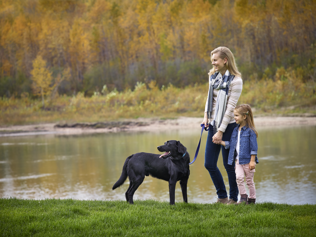 A mother and daughter walk their black lab dog beside the river, with fall colored trees in the background.